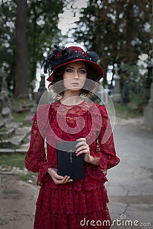 Woman in a vintage red dress and a black hat stands in a cemetery among the graves and holds a bible Stock Photo