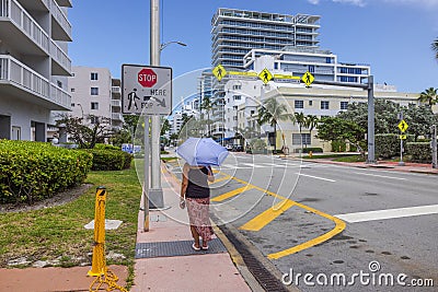 Woman walks down Collins Avenue on scorching, sunny day, shielding herself from sun's rays with UV-protective umbrella. Editorial Stock Photo