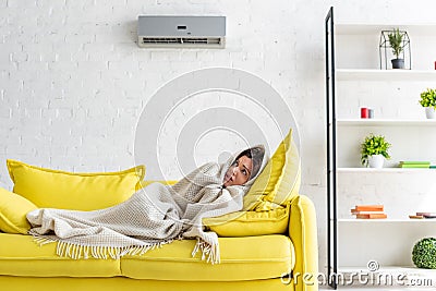 Woman warming with blanket while lying on yellow sofa under air conditioner at home Stock Photo