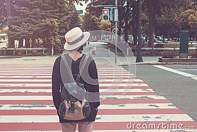 Woman wear white weave hat and black overcoat. She standing on footpath and waiting for crossing the road. Stock Photo