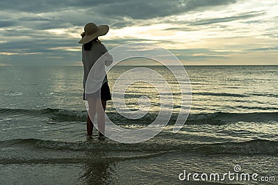 Woman wearing a hat walks alone on an empty sandy beach at sunset. A lonely and depressed young woman stands on the sand of the be Stock Photo