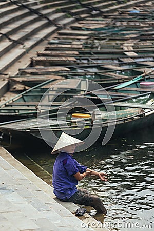 Woman wearing purple shirt, conical hat carry wash her feet in the river with empty rowing boats in the background at Trang An Editorial Stock Photo