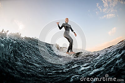 woman in wetsuit wakesurfing on the board and riding down the river wave and show hand gesture Stock Photo