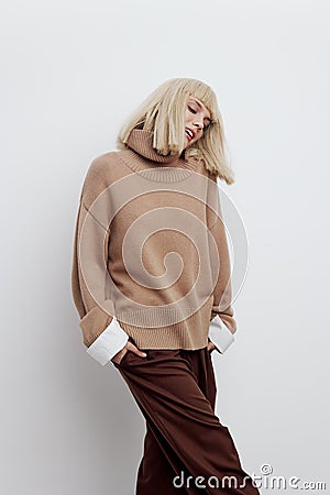 a woman on a white background in a beige sweater and brown trousers with fine blond hair stands and smiles with her Stock Photo