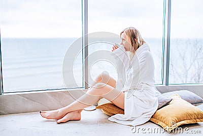 Woman in a white bathrobe enjoying sea view next to big window with cup of coffee. Panoramic window. Floor covering, Vacation in a Stock Photo