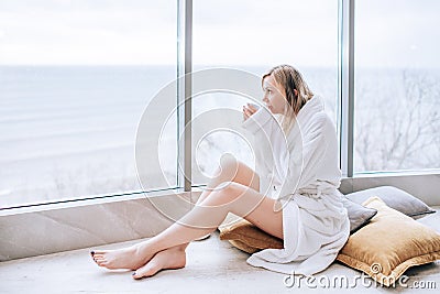 Woman in a white bathrobe enjoying sea view next to big window with cup of coffee. Panoramic window. Floor covering, Vacation Stock Photo