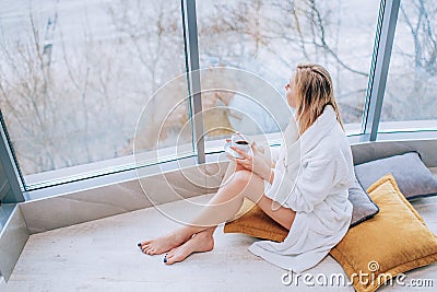 Woman in a white bathrobe enjoying sea view next to big window with cup of coffee. Panoramic window. Floor covering, Vacation Stock Photo