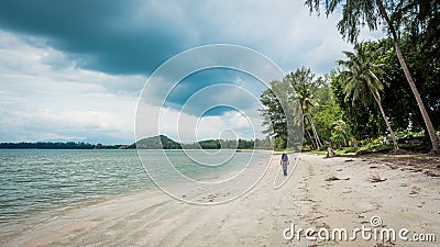 Woman in white fedora hat and sarong sown the beach with palm trees and storm clouds in the background Stock Photo