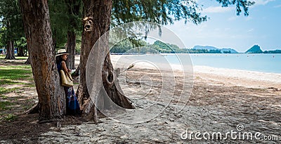 Woman with white fedora and sarong leaning against tree at the beach looking at the sea Stock Photo
