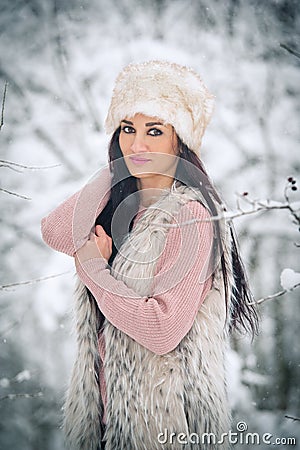 Woman with white fur cap and sheepskin smiling enjoying the winter scenery in forest. Side view of happy brunette girl posing Stock Photo