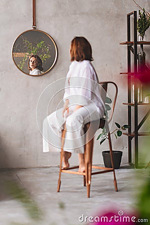 a woman in white on a high chair looks at the reflection in a round mirror. Stock Photo