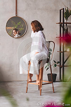 a woman in white on a high chair looks at the reflection in a round mirror. Stock Photo