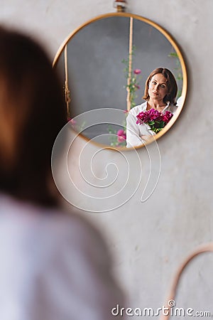 a woman in white with a peonies looks at the reflection in a round mirror. Stock Photo