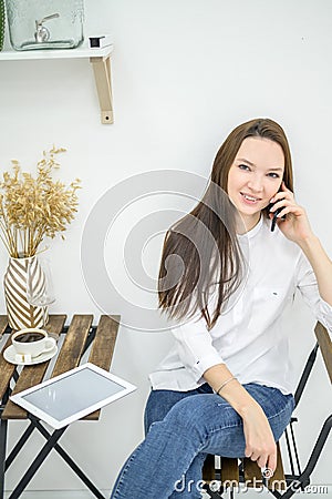 A woman in a white shirt and jeans sits in a cafe and drinks coffee. Female office worker at the lunch break talking on Stock Photo