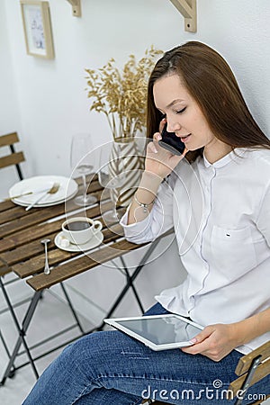 A woman in a white shirt and jeans sits in a cafe with a tablet in her hands. Female office worker at the lunch break Stock Photo