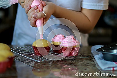 woman in white shirt spreading pink buttercream frosting on three cupcakes, blurred background Stock Photo