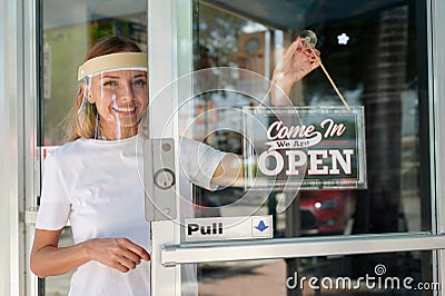 Smiling woman in white shirt wearing face shield hanging open sign for storefront business on the door Stock Photo