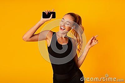 Woman In Wireless Earbuds Holding Smartphone And Dancing, Studio Shot Stock Photo