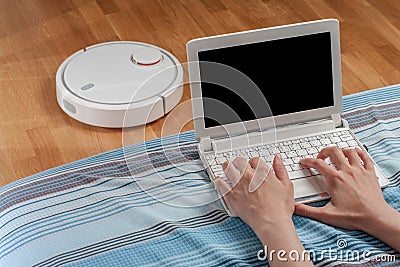 woman works at computer in living room while robot vacuum cleaner vacuuming laminate floor. automation homework, facilitation rout Stock Photo