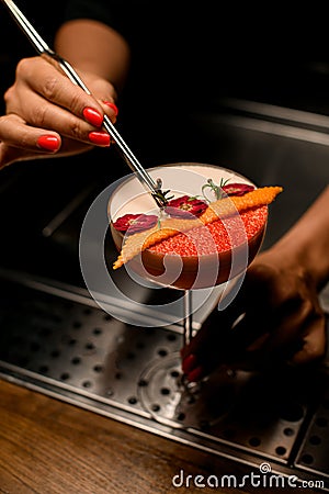 woman& x27;s hand holds tweezers with twig and decorates glass with frothy drink and flowers Stock Photo