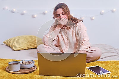 A woman yawns while sitting at her laptop in the evening room. Red-haired girl working with food on the bed Stock Photo
