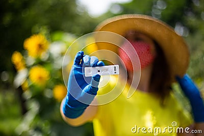 Woman in yellow top wearing a red facemask and hat holding a coronavirus rapid test in front of sunflowers bokeh Editorial Stock Photo