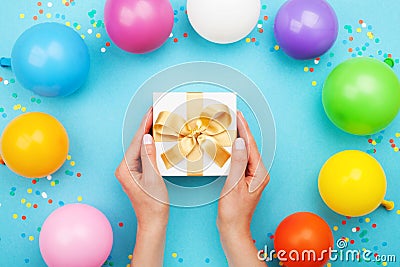 Womans hands holding gift or present box on blue table decorated colorful balloons and confetti. Flat lay style. Stock Photo