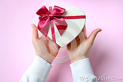 Womans hands holding gift or present heart shaped box decorated big red bow on pink table top view. Stock Photo