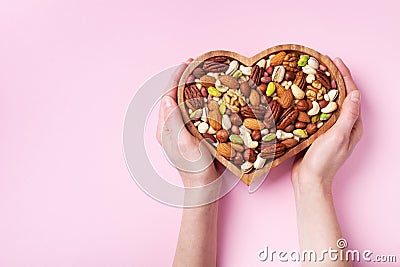 Womans hands holding heart shaped bowl with mixed nuts on pink table top view. Healthy food and snack. Flat lay Stock Photo