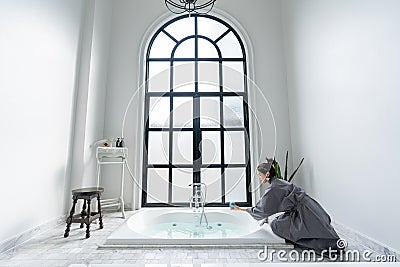 Women in bathrobe while carryin bathbomb in nice design jacuzzi bathtub with high transparant window in natural light setting Stock Photo