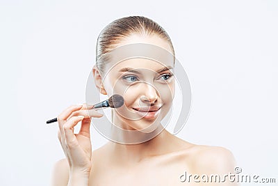 Women, cosmetology concept. Tender healthy European woman gives makeup lesson, applies cosmetic with beauty brush, looks aside Stock Photo