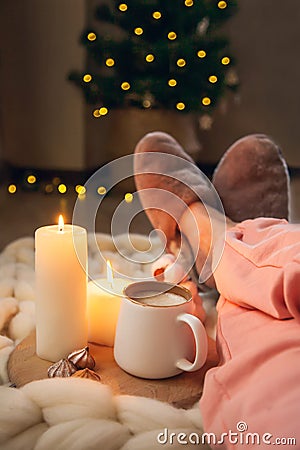 Women`s feet in home fur slippers, cup of cappuccino, candles on blanket of thick yarn and Christmas tree with lights Stock Photo