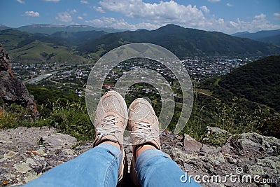 Women's legs in blue jeans and light sneakers against the backdrop of mountains, the old city and the blue sky. Stock Photo