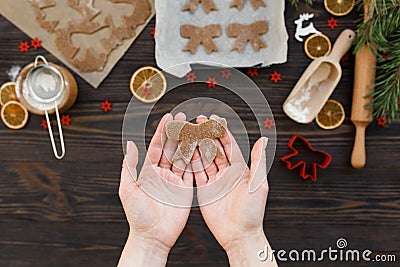 On women`s palms is Christmas cookie figure in a bow shape. Kitchen wood table with cutters, rolling pin, dough, flour Stock Photo
