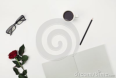 Womens working space with cup of coffee, pencil, empty notebook, eyeglasses and rose flower on white table from above. Flat lay. Stock Photo