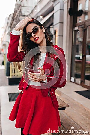 Wonderful girl in red attire looking to camera through dark glasses. Outdoor photo of ecstatic brunette caucasian woman Stock Photo