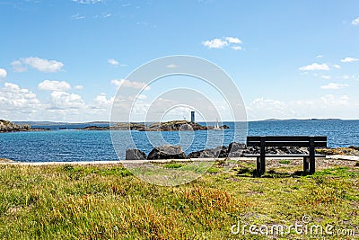 Wooden bench on the coastal shoreline in Inishbofin with the lighthouse on Gun Rock and a sailboat in the background Stock Photo