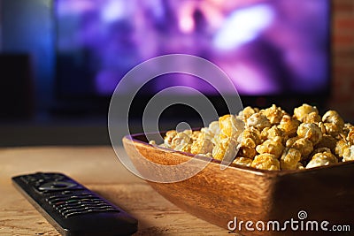 A wooden bowl of popcorn and remote control in the background the TV works. Evening cozy watching a movie or TV series at home Stock Photo