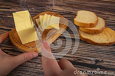 Wooden butter dish with a piece of butter and slices of cheese and fresh wheat toast on a wooden background. Someone cuts off a Stock Photo