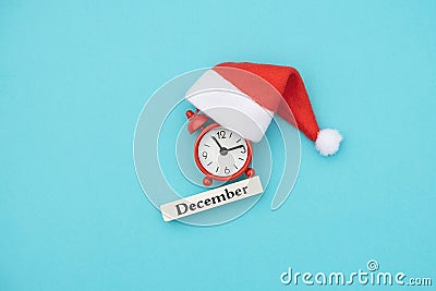 Wooden calendar winter month December, red alarm clock and Santa hat on blue paper background. Concept Hello December, xmas. Stock Photo