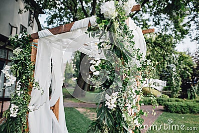 Wooden ceremony arch decoretade by white cloth, flowers and greenery standing in bright garden for wedding ceremony. Decor. Stock Photo