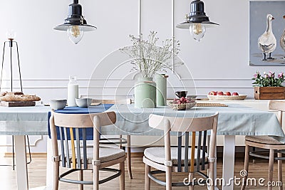 Wooden chairs at table with flowers and food in white cottage dining room interior with lamps and poster. Real photo Stock Photo