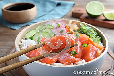 Wooden chopsticks with piece of salmon over delicious poke bowl on table, closeup Stock Photo