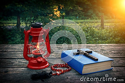 Wooden christian cross on bible, burning kerosene lamp and prayer beads on the old table. Salvation of soul and expiation of sins. Stock Photo