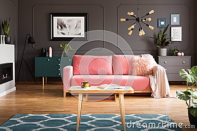 Wooden coffee table with tea cup and open book standing on carpet in dark living room interior with powder pink lounge with fur c Stock Photo