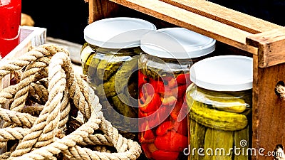 Wooden crate with glass jars with pickled red bell peppers and pickled cucumbers (pickles) isolated. Stock Photo