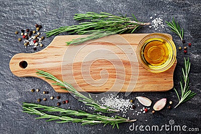 Wooden cutting board, olive oil, rosemary plant, salt, garlic and pepper on black table from above for food cooking background or Stock Photo