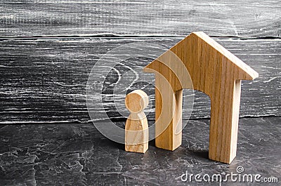 A wooden figure of a man stands near a wooden house on a gray concrete background. Concept of real estate, renting and buying Stock Photo