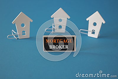 Wooden houses sits next to a wooden black board with the red word mortgage broker. Beautiful blue background. Business and Stock Photo