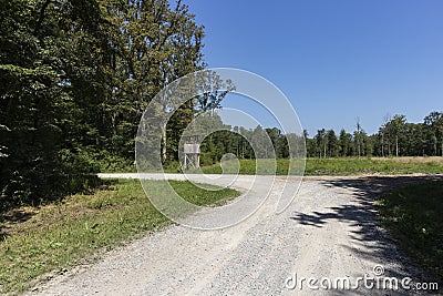 Wooden hunting post standing next to gravel path on the forest edge where wildlife go to graze the grass on the meadow behind near Stock Photo